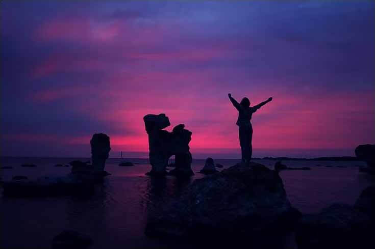 Sunset photo of a woman standing silhouetted against the blue ocean and pink and purple sky.